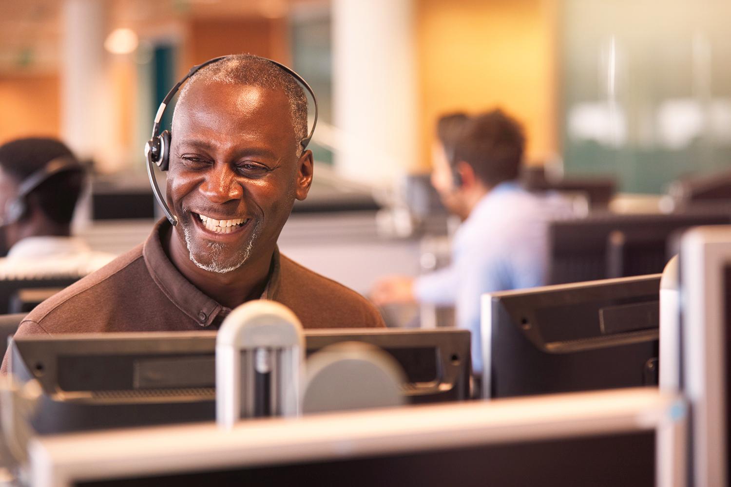 A claims professional wearing a headset smiles at their workstation while colleagues work nearby.